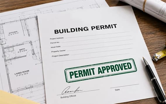 Building permit form stamped Permit Approved on a desk with architectural floor plans, a pen, and a tape measure.