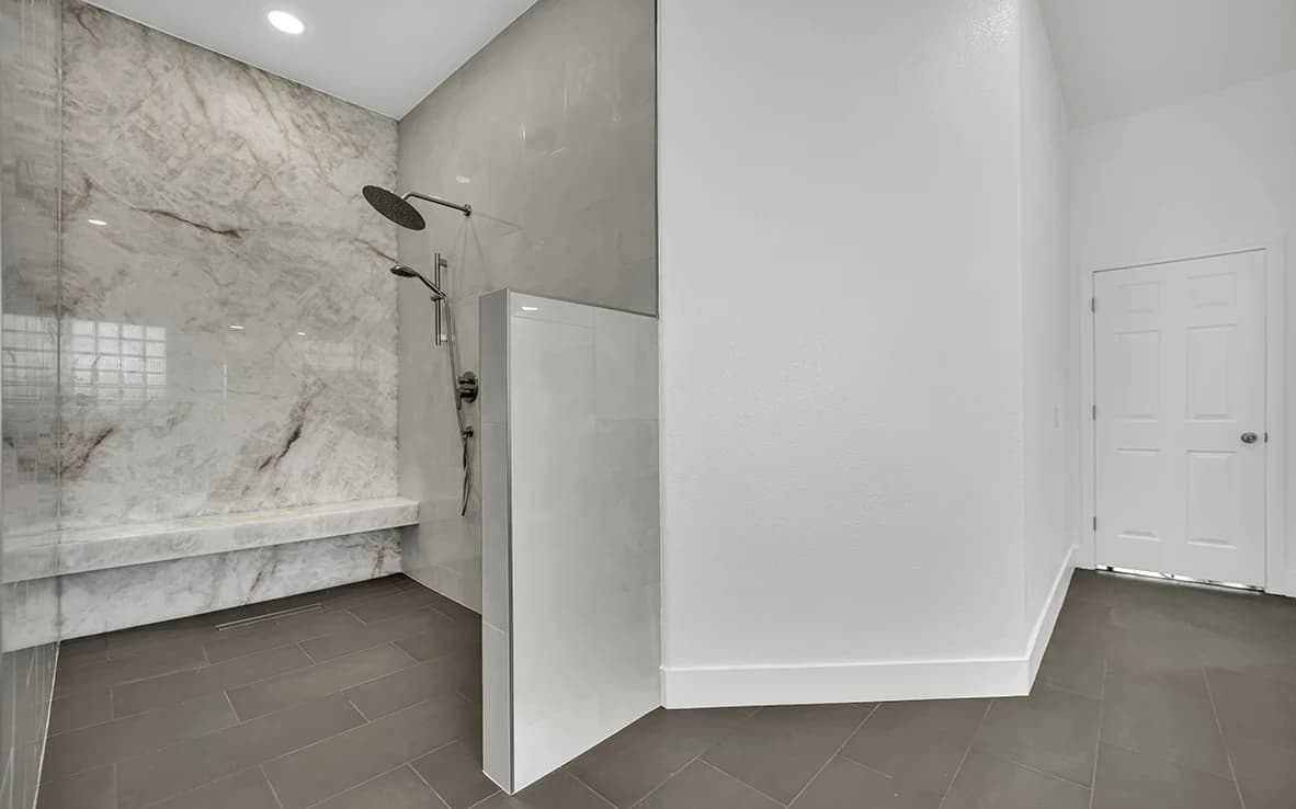 Wide view of an expanded curbless shower with onyx slab wall, partial glass, and continuous charcoal gray bathroom floor tile