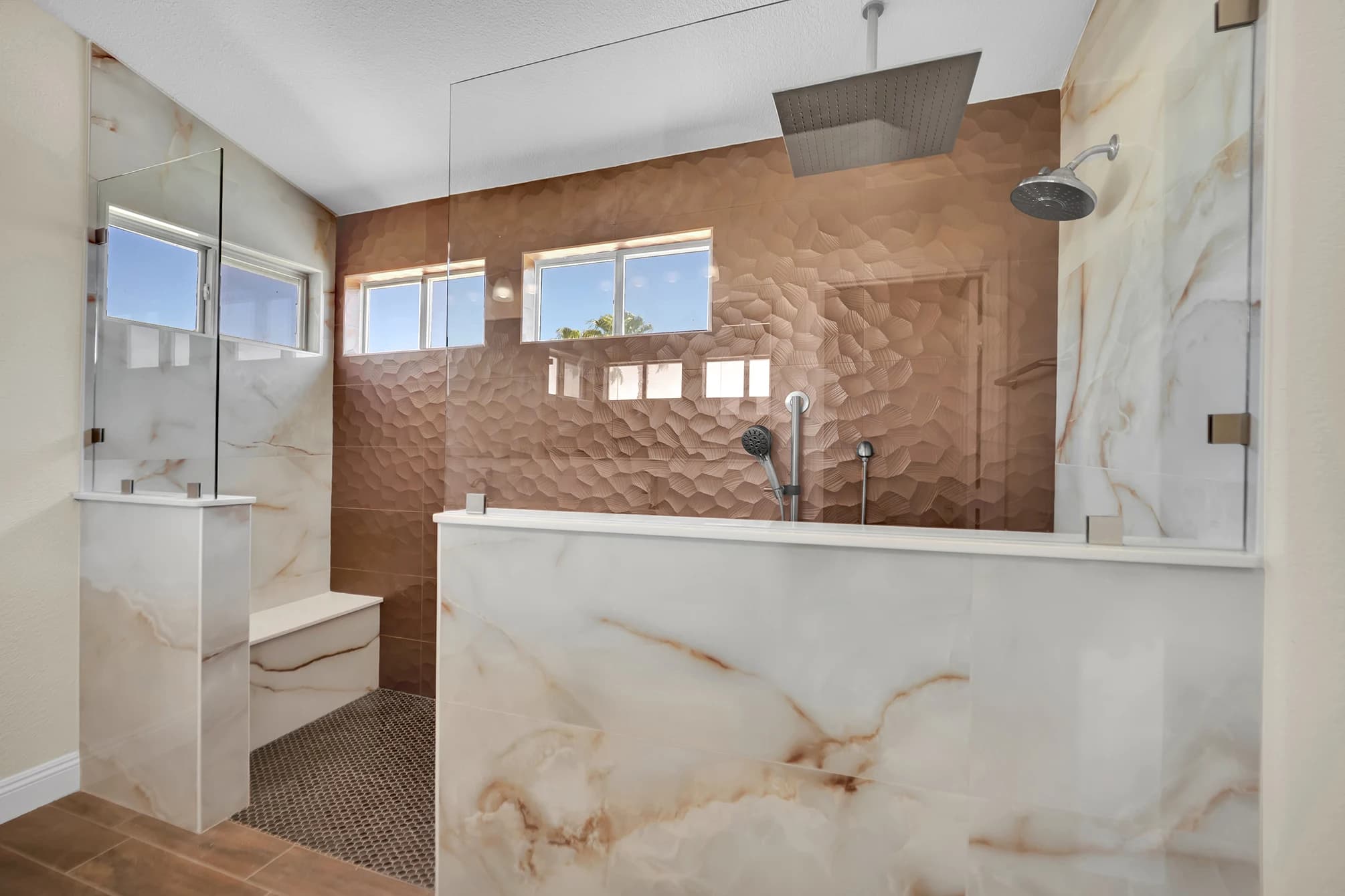 Centennial Hills curbless shower with copper-tone accent tile, cream veined pony wall and ceiling-mounted Kohler rainhead.