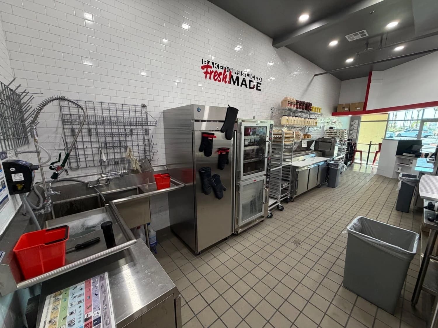 Stainless steel three-compartment sink, commercial oven and preparation equipment along a tiled restaurant kitchen.