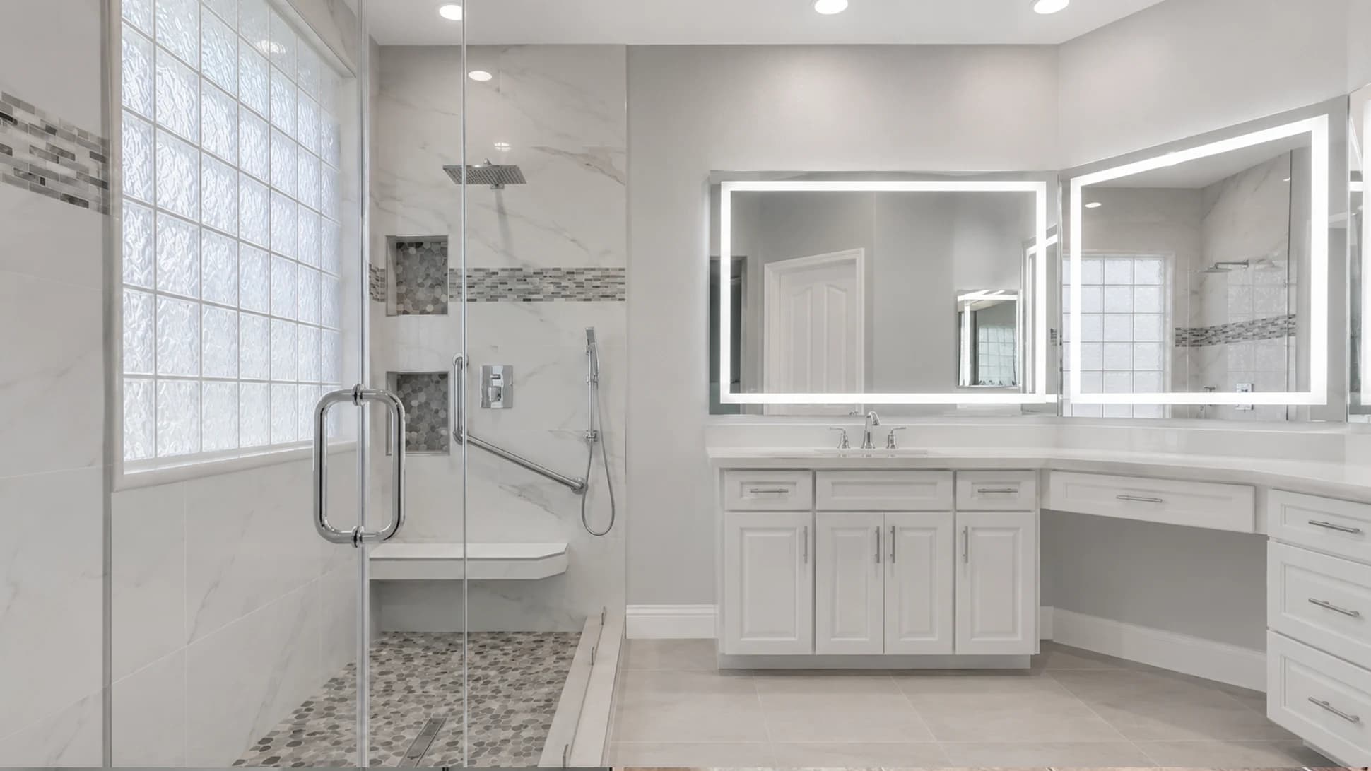 Finished bathroom after the curbless walk-in shower conversion with fixed glass, pebble floor, grab bars, white vanity, and lighted mirrors