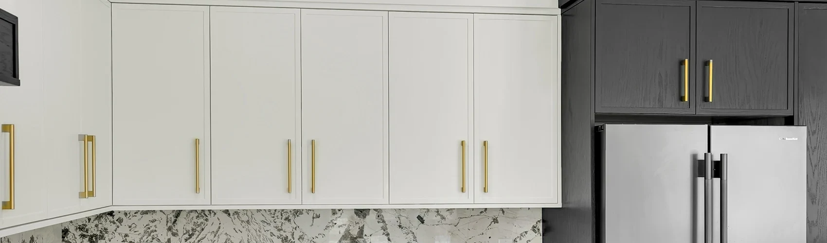 Kitchen cabinet wall with white shaker cabinets, dark refrigerator surround, brass hardware, and slab backsplash