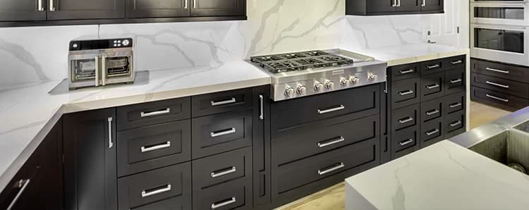 Completed kitchen with dark base cabinets, white slab backsplash, gas range, white upper cabinets, and light stone counters