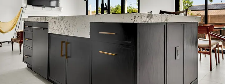 End of dark kitchen island with storage cabinets, waterfall-style stone counter, and dining seating nearby