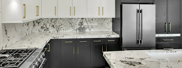 Range wall with slab backsplash, white upper cabinets, dark lower cabinets, stainless refrigerator, and stone island edge