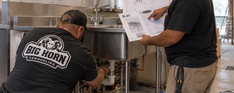 Two Big Horn Remodeling crew members work below a commercial sink while reviewing an R-01 equipment specification sheet.