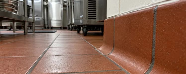 Red quarry tile floor meeting a curved tile cove base along a white commercial kitchen wall.