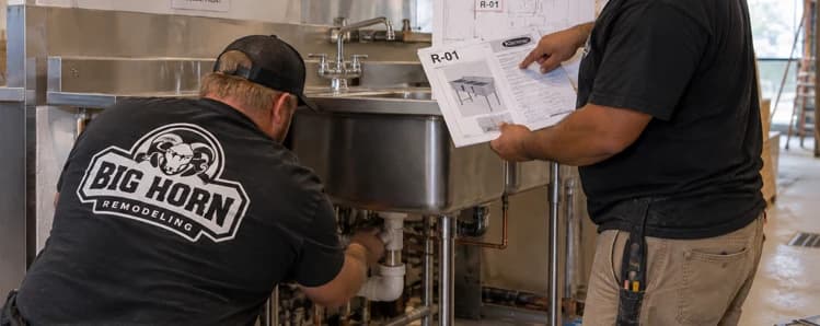 Two Big Horn Remodeling crew members inspect a commercial sink and plumbing while reviewing an R-01 equipment specification sheet.