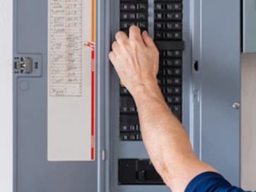 Open electrical panel with circuit labels and a hand checking the breakers.