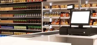Retail checkout counter with a point-of-sale monitor, cash drawer and stocked shelves behind it.