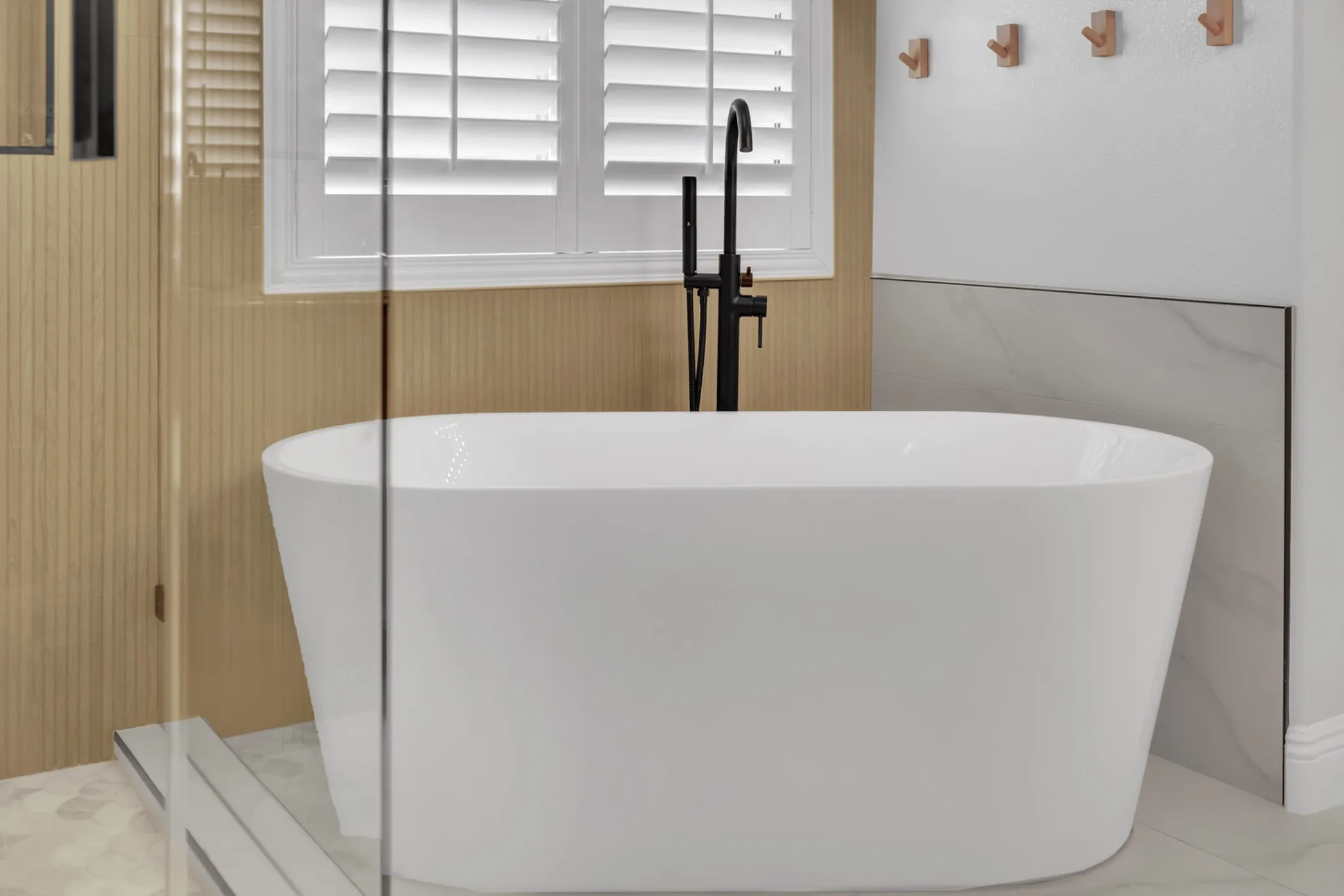 Bathroom remodel detail showing wood shelf, robe hooks, plantation shutters, matte black freestanding tub filler, and marble-look wall tile
