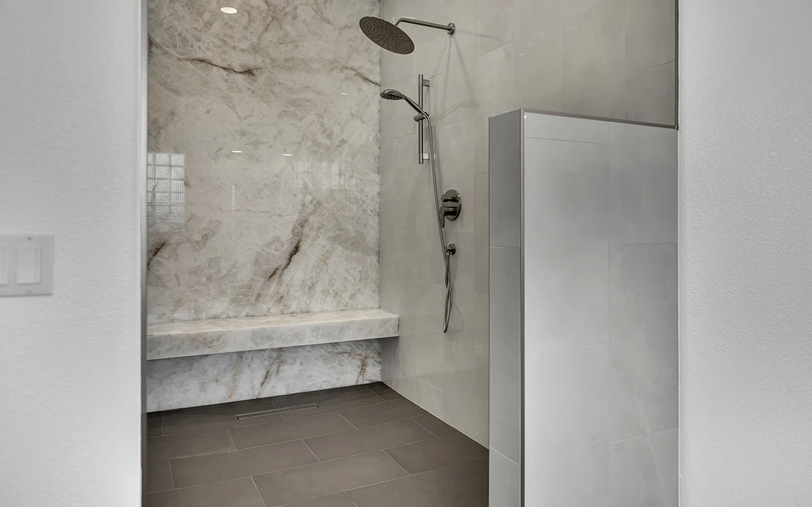 Curbless primary bathroom remodel with Honey Amber Onyx slab shower wall, bench, linear drain, rain head, handheld wand, and charcoal gray tile floor