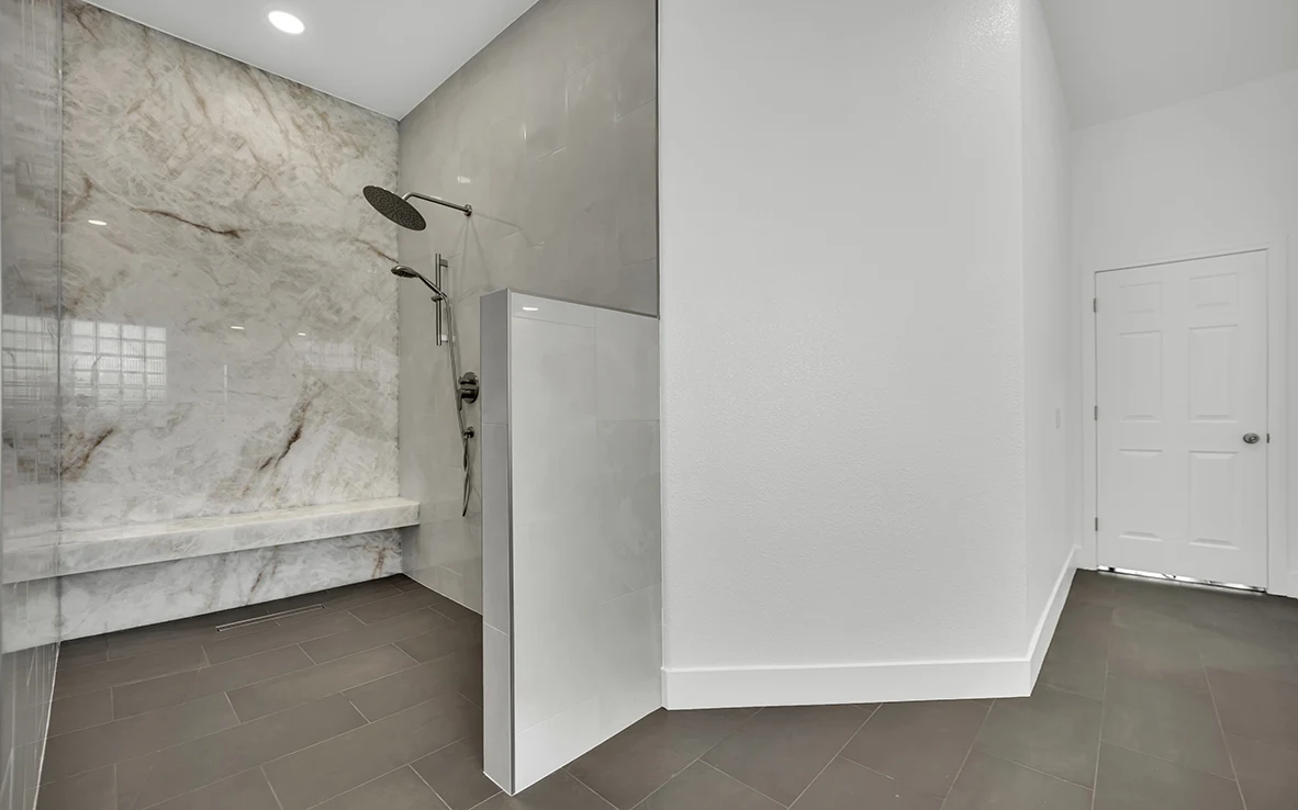 Wide view of an expanded curbless shower with onyx slab wall, partial glass, and continuous charcoal gray bathroom floor tile