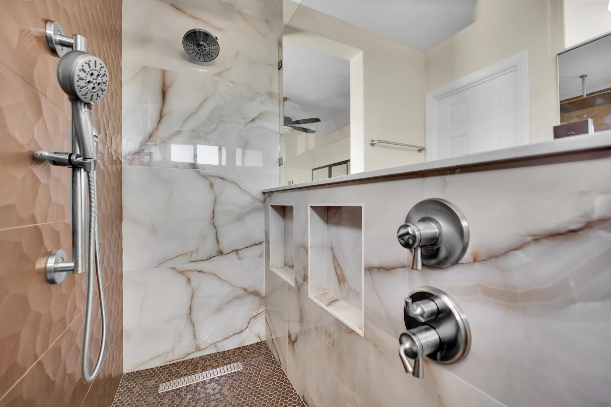 Two recessed shower niches beside Kohler controls, with a handheld shower and linear drain in the penny mosaic floor.