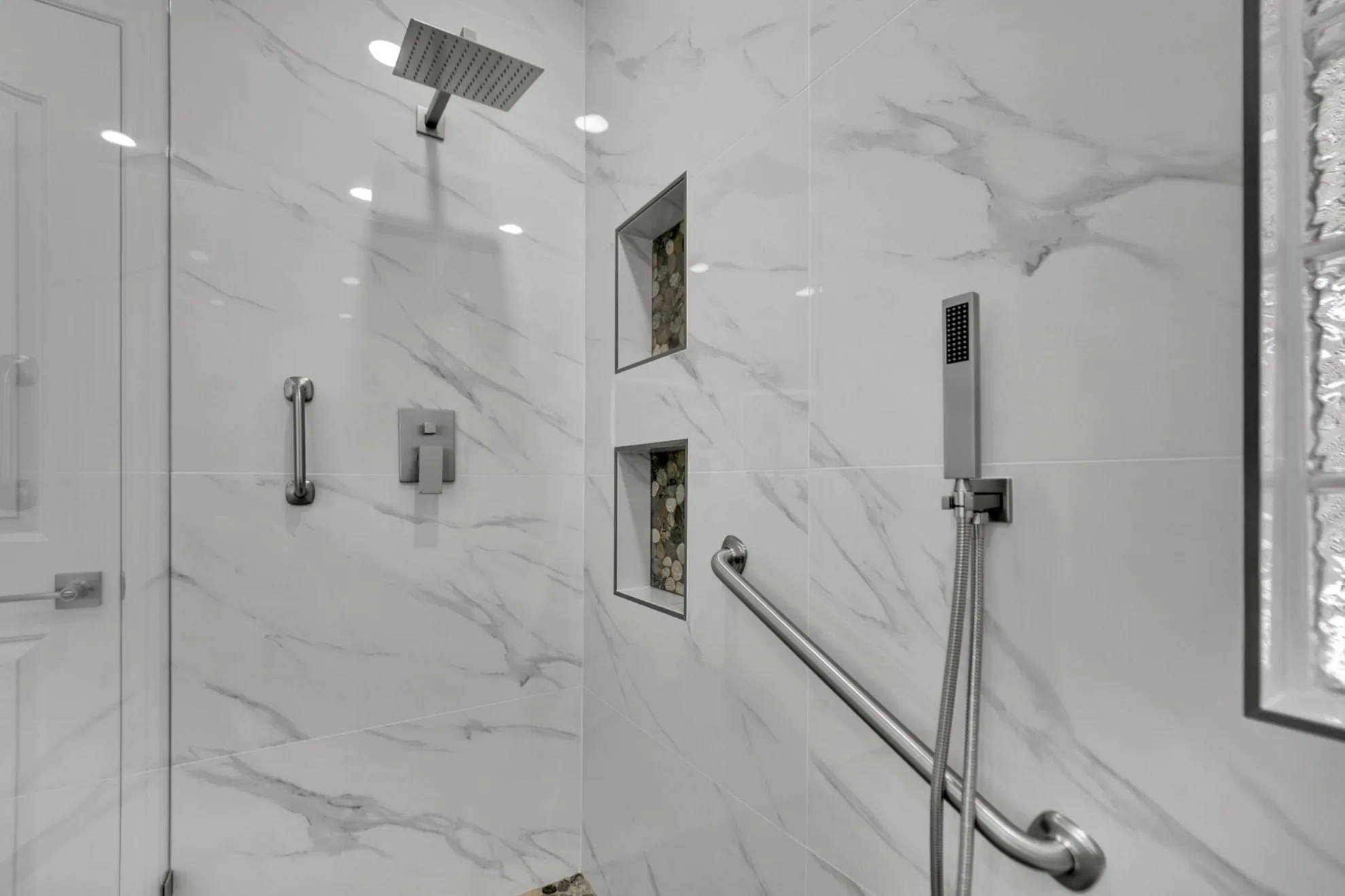 Close-up of curbless shower with built-in niches, handheld shower wand, grab bar, and marble-look porcelain tile