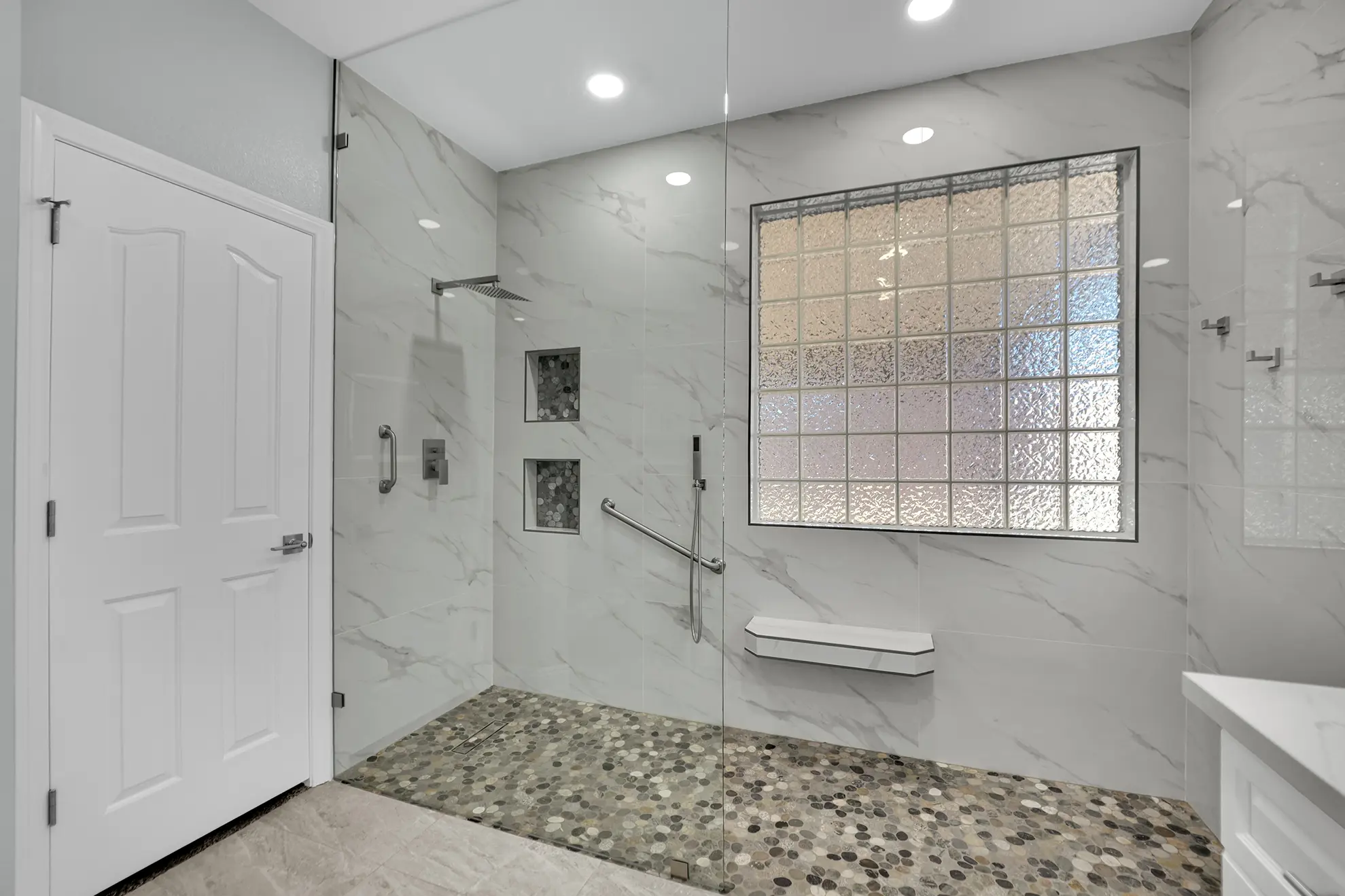 Accessible curbless shower with fixed glass, pebble tile floor, grab bars, glass block window, and floating bench