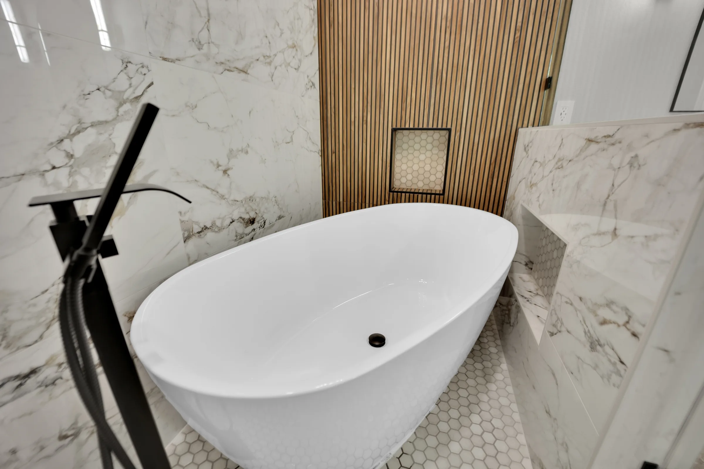White freestanding tub on hexagonal floor tile beside a niche and marble-look pony wall.